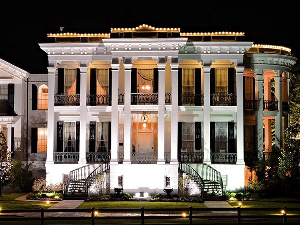 A grand white mansion illuminated at night, featuring multiple columns, large windows, and decorative railings, surrounded by landscaped gardens and a pathway.
