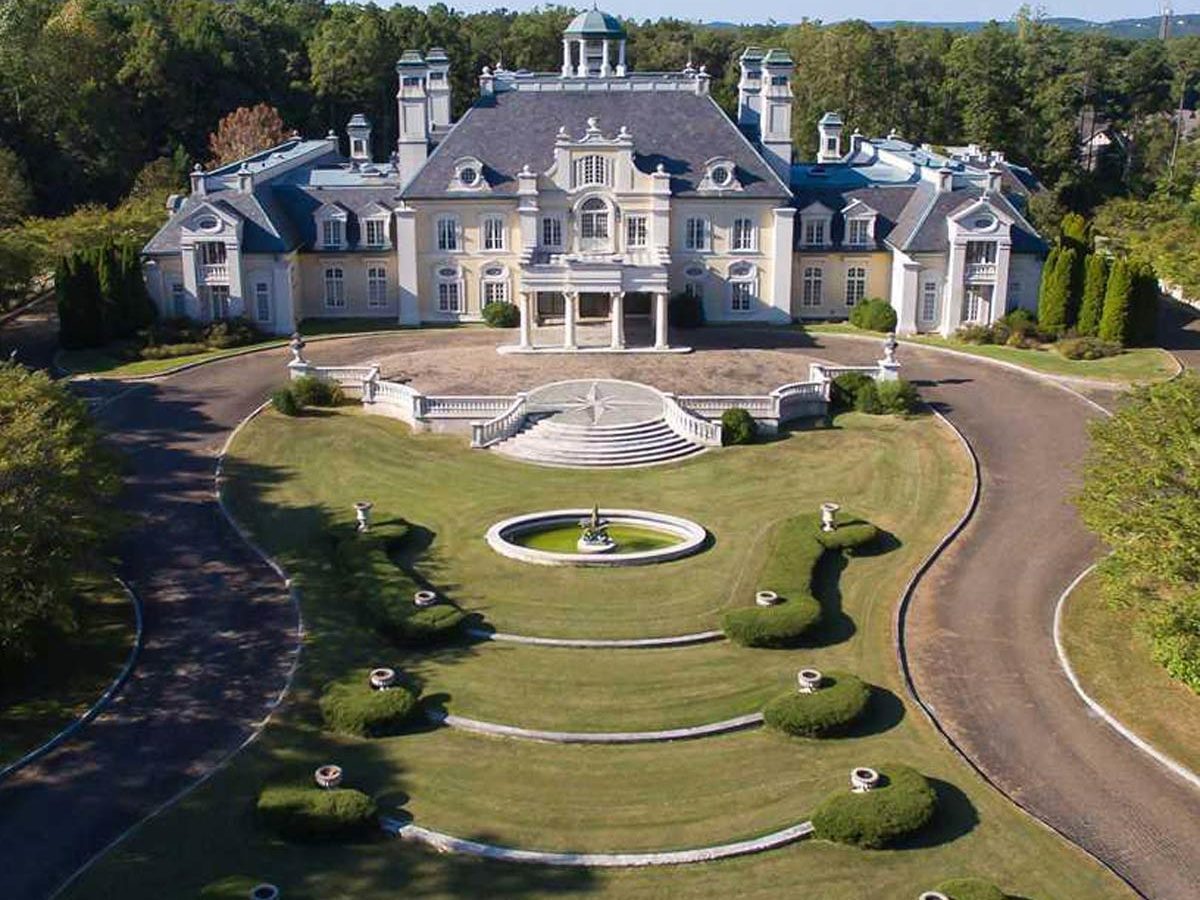 A grand mansion stands prominently, surrounded by manicured lawns and ornamental bushes, with a circular fountain at the center of the landscaped garden.