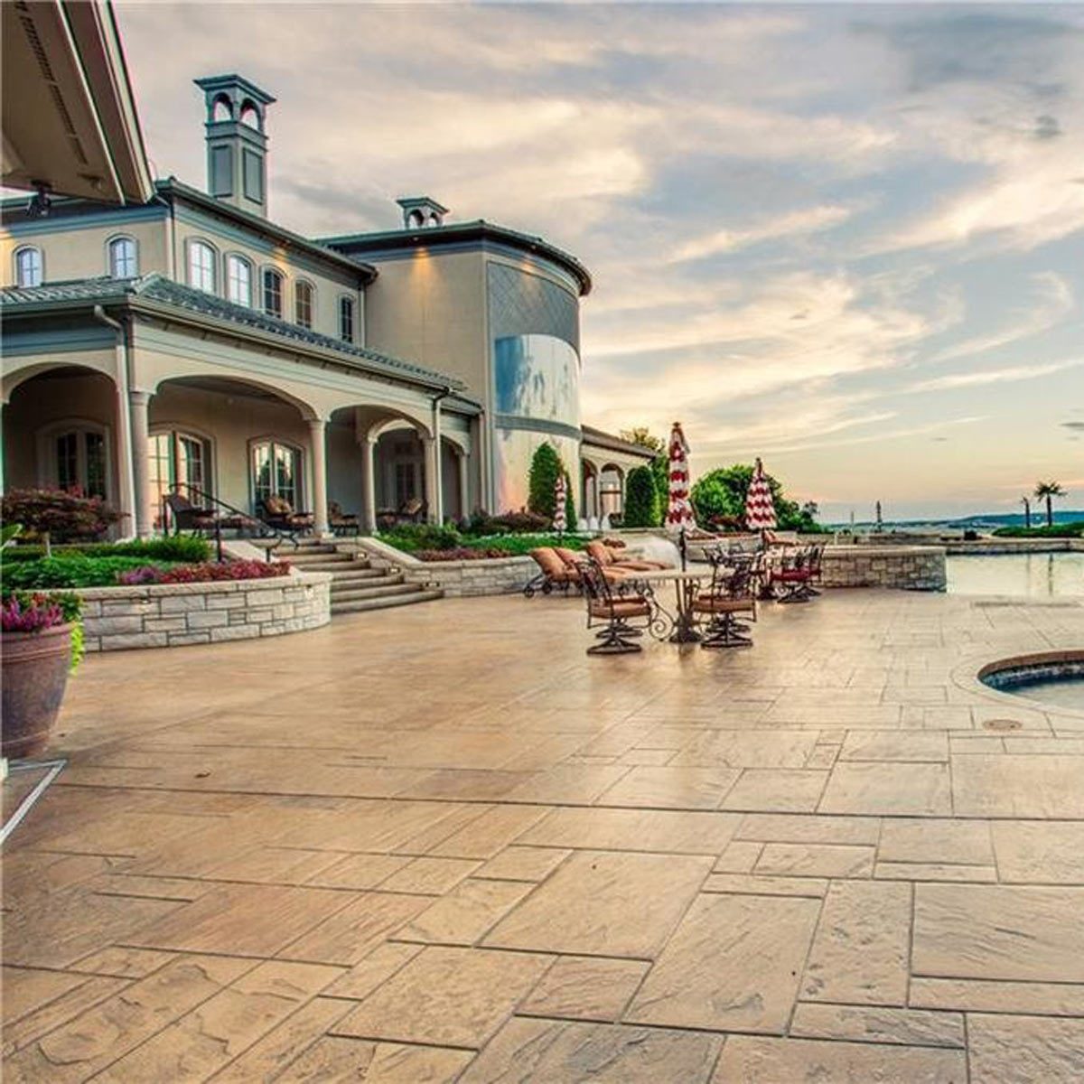 A luxurious patio with decorative chairs and potted plants, leading to a large home overlooking a scenic view under a partly cloudy sky.
