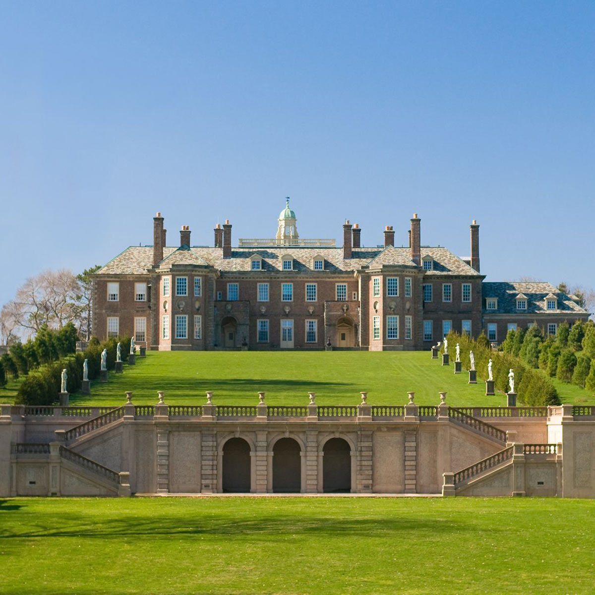 A large, elegant mansion stands on a green lawn, with statues lining the pathways and a clear blue sky overhead.