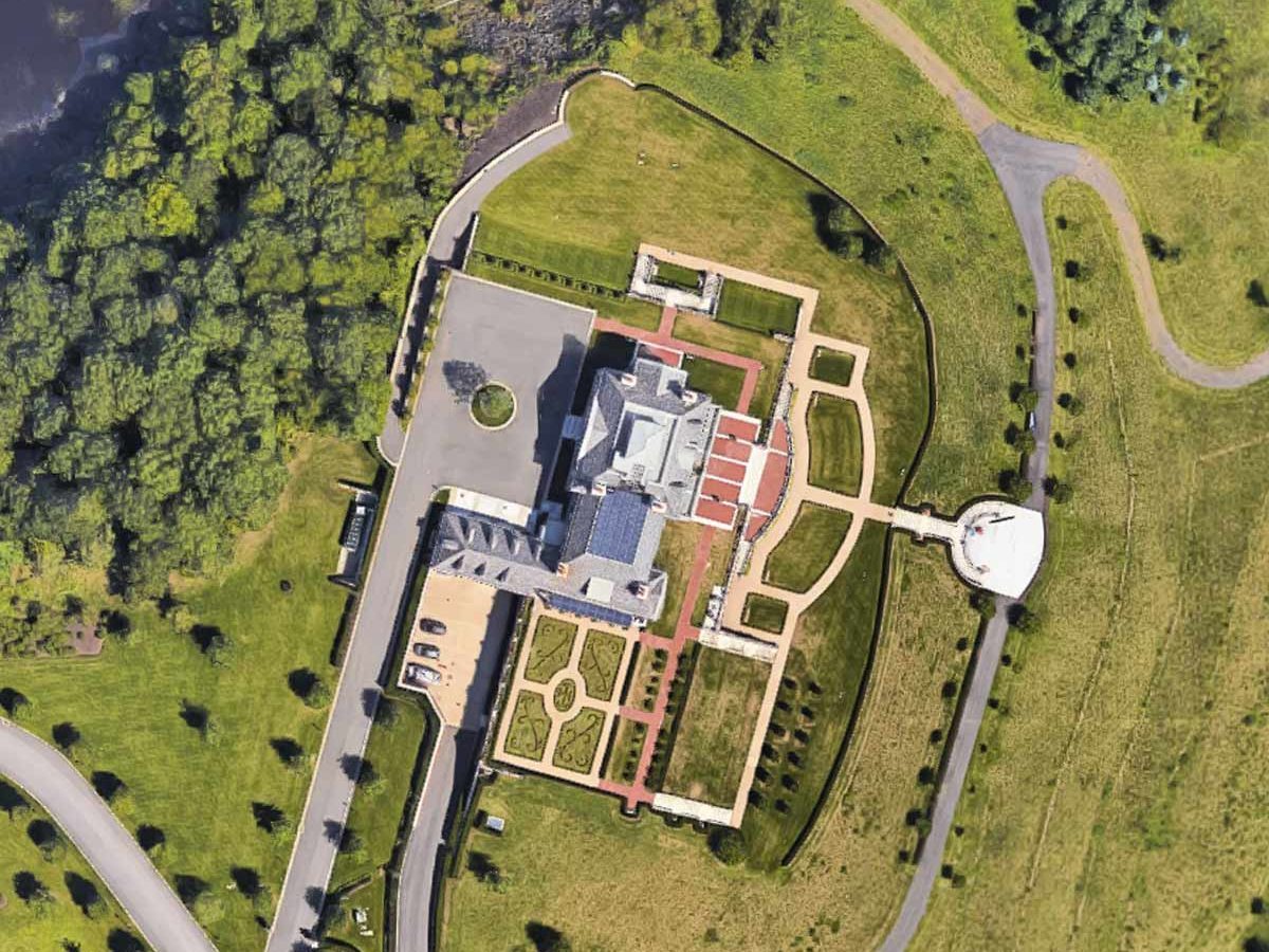 A large residential building is positioned amidst manicured lawns and gardens, with surrounding trees and a winding road leading to the structure.