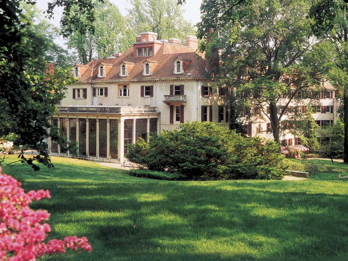 A large, elegant house with multiple rooflines stands amidst lush green grass and trees. Bright pink flowers frame the scene, adding color to the landscape.