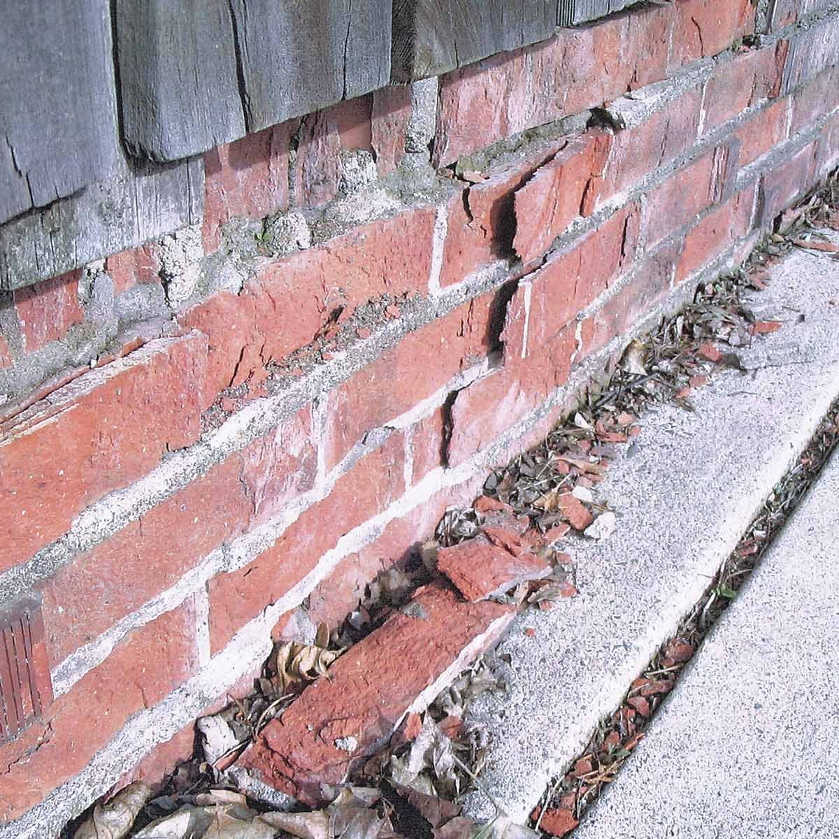 A cracked brick wall reveals loose bricks, with debris and dried leaves scattered along a concrete edge in an outdoor setting.
