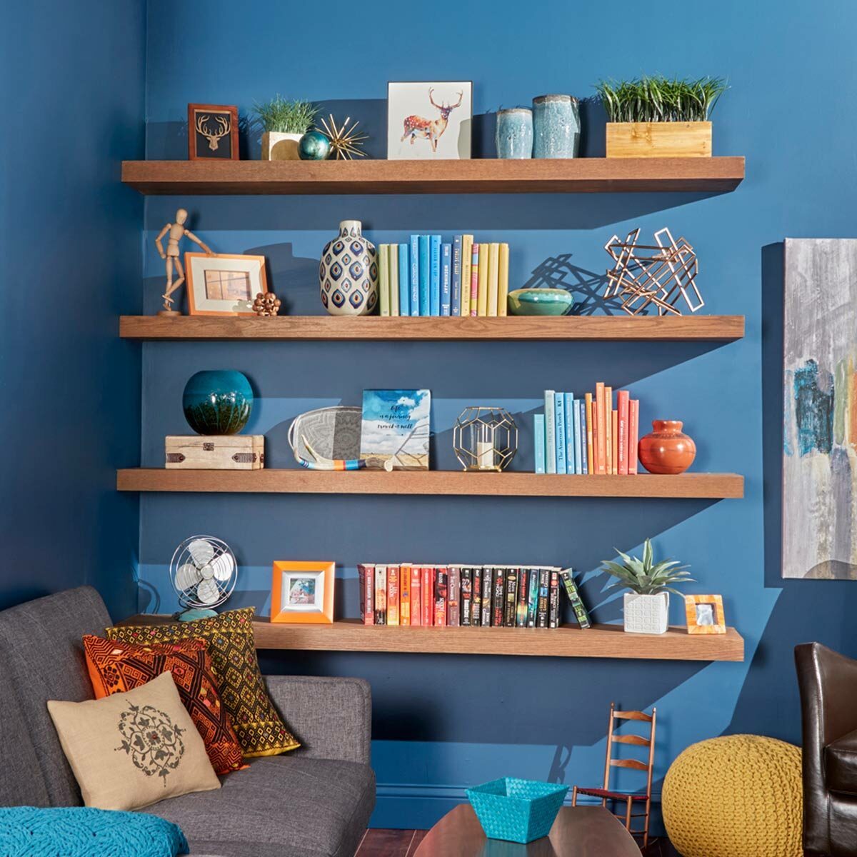 Wooden shelves display colorful books and decorative items against a blue wall, while a gray sofa with patterned cushions and a small table complete the cozy room.