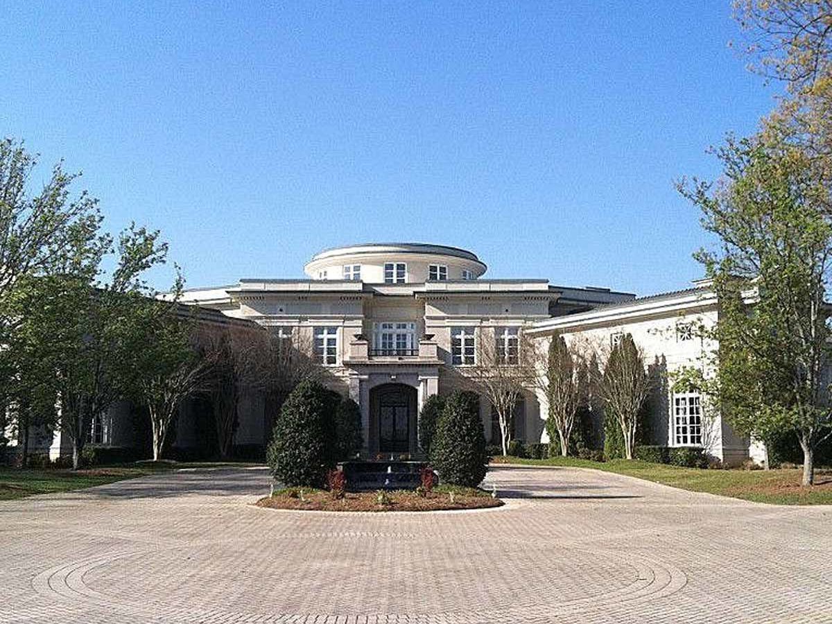 A large, elegant mansion stands in a paved courtyard, featuring lush greenery and a clear blue sky in the background.