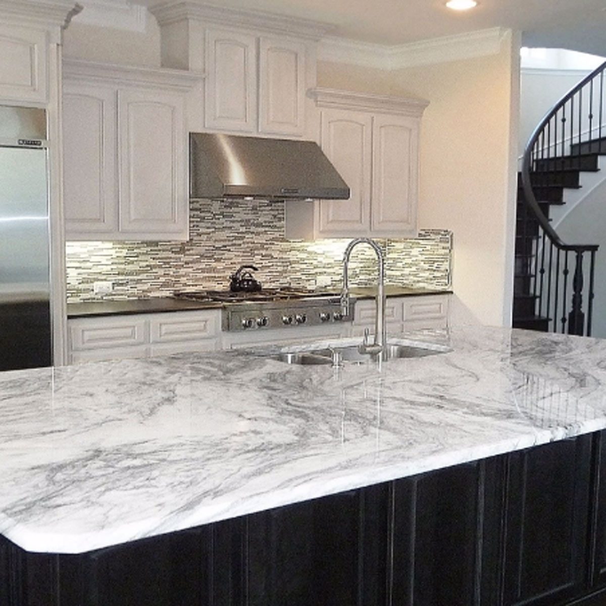 A marble kitchen island with a sink showcases stainless steel appliances and a patterned backsplash, while a spiral staircase is visible in the background.