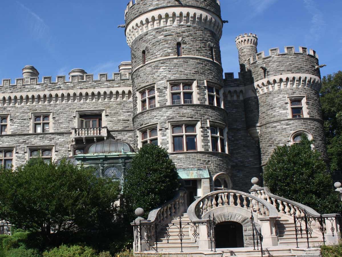 A stone castle features turrets and large windows, surrounded by green shrubbery and stairs leading to an entrance, under a clear blue sky.