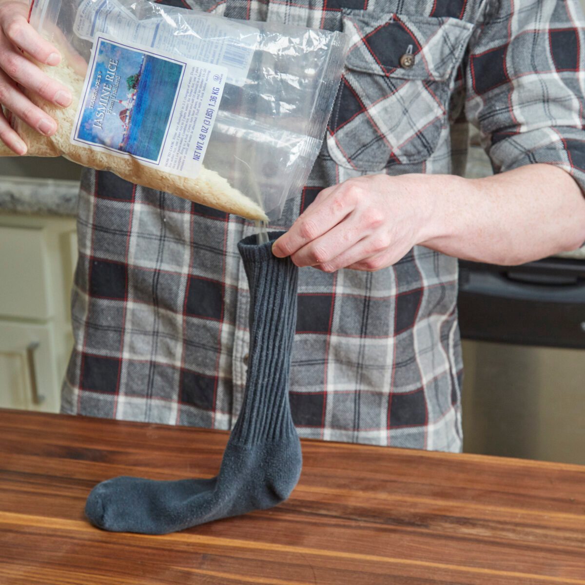 A person holds a bag of rice, pouring it into a dark sock placed upright on a wooden countertop in a kitchen setting.