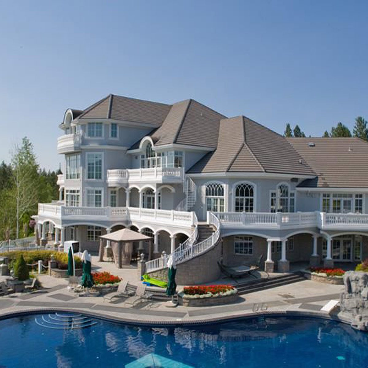 A large, elegant mansion features multiple balconies and a grand staircase, beside a landscaped pool area with sun loungers and vibrant flowers. The sky is clear.