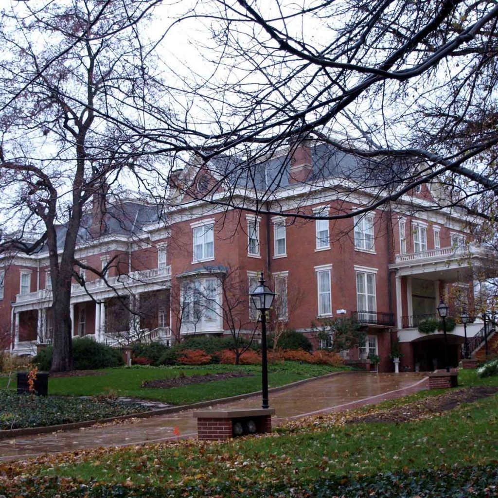 A large brick mansion stands prominently, surrounded by bare trees and autumn foliage, as rain-soaked paths lead up to its entrance.