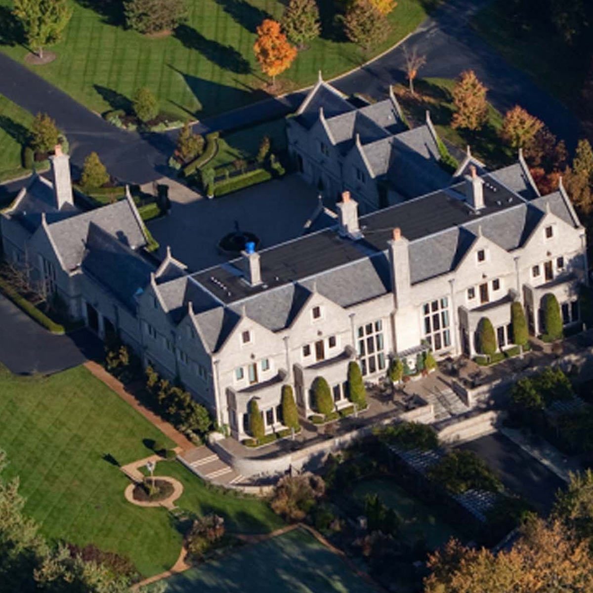 A large, elegant mansion with a landscaped garden is surrounded by trees and a paved driveway, highlighting its expansive architecture against a clear backdrop.