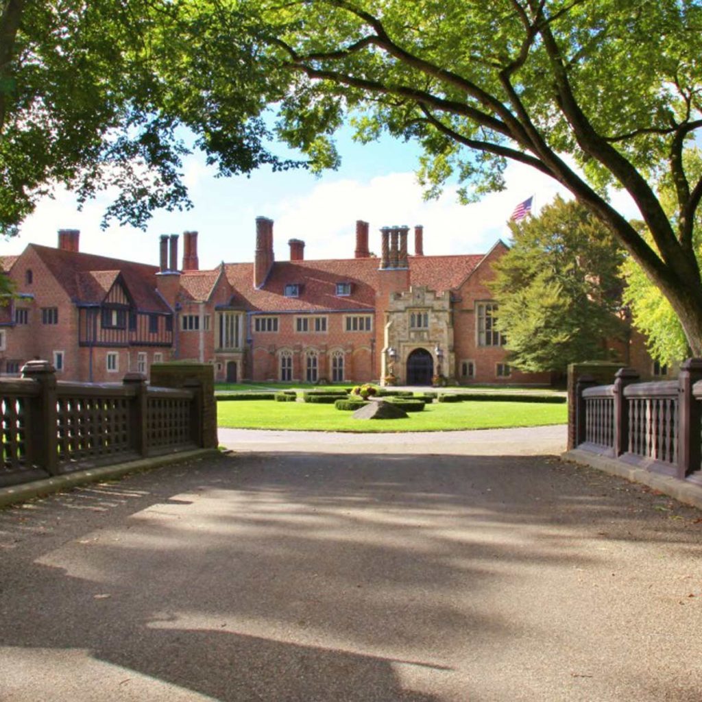 A large, elegant mansion stands prominently, surrounded by manicured lawns and trees, inviting visitors through a decorative gate along a paved pathway.