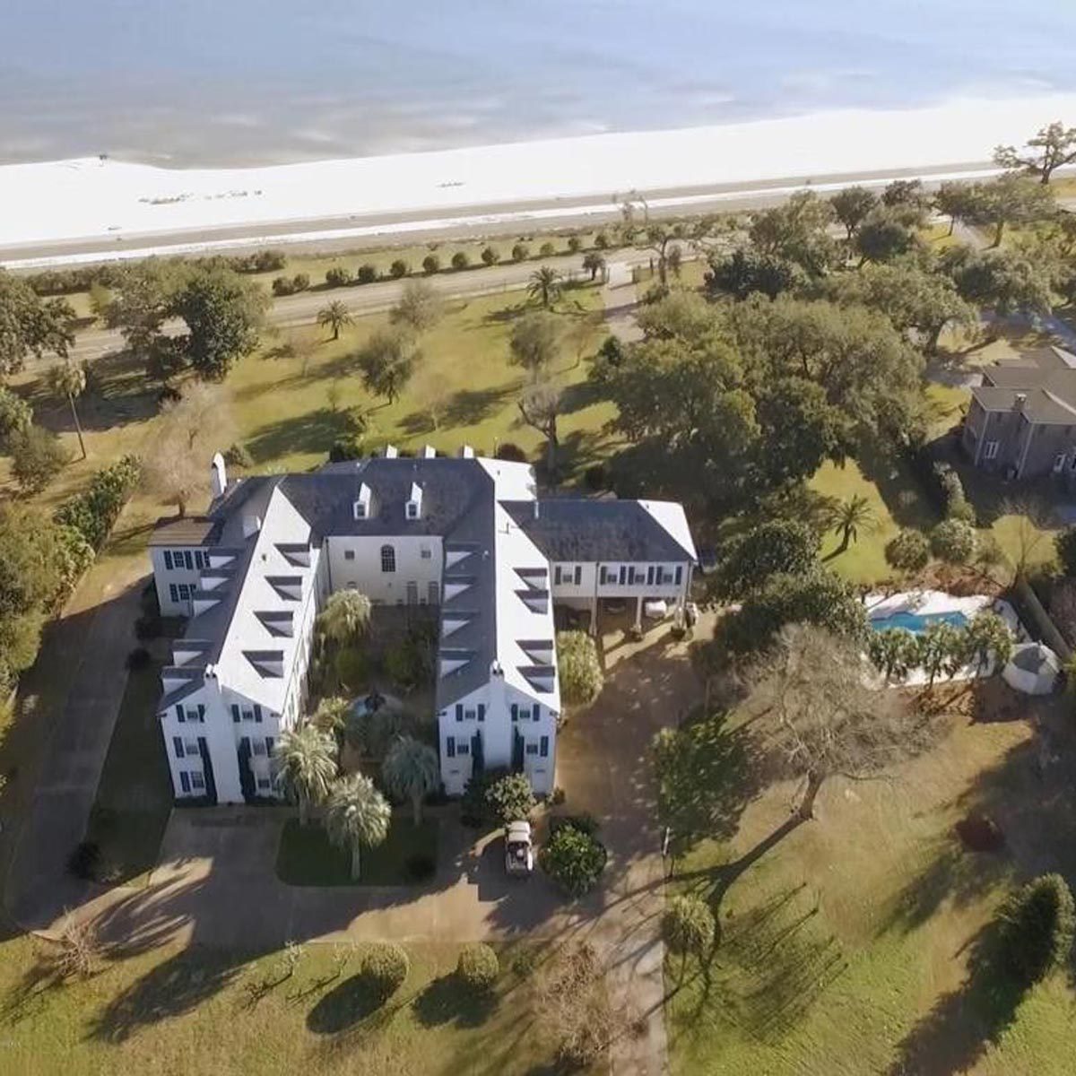 A large, multi-story house with a pool is surrounded by greenery, located near a sandy beach and road under a clear sky.