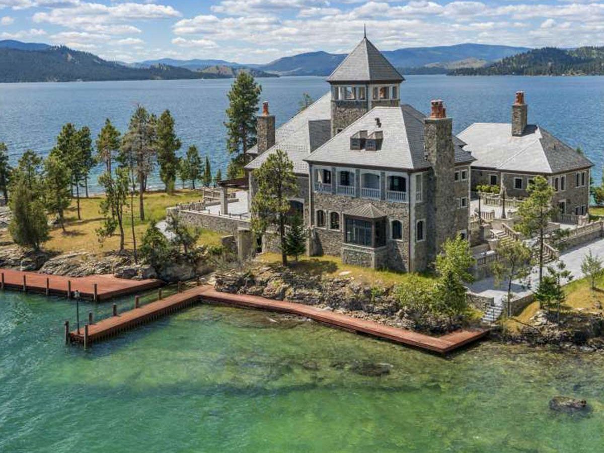 A grand stone mansion sits by a clear lake, surrounded by trees and mountains, with a wooden dock extending into the water under a blue sky.