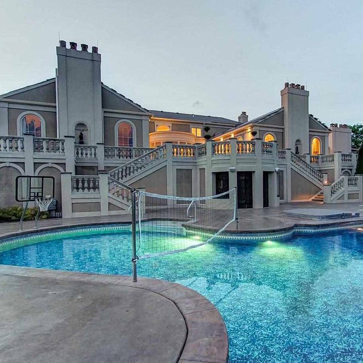 A large house features a pool with illuminated water, a volleyball net, and basketball hoop, surrounded by staircases and decorative railings in a twilight setting.