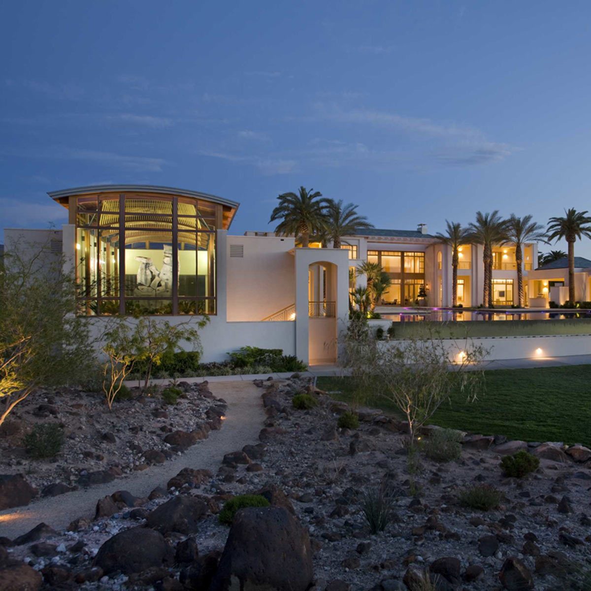 A modern home with large glass windows is illuminated at dusk, surrounded by landscaped stone pathways and palm trees in a serene outdoor setting.