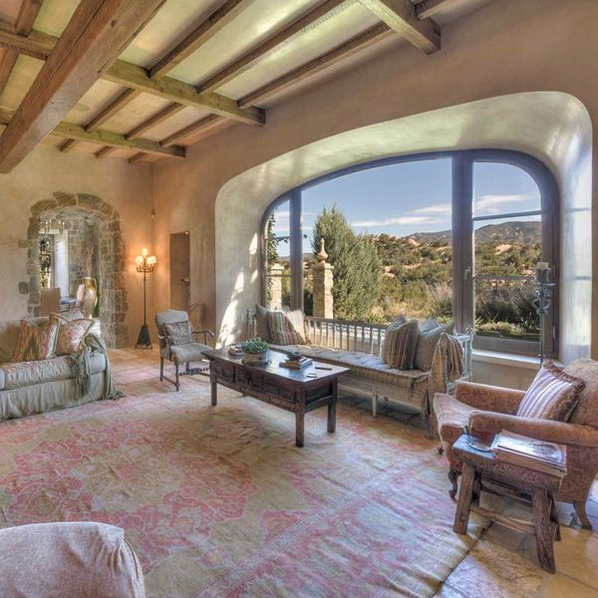 A cozy living room features a wooden table surrounded by upholstered seating. Large windows reveal a scenic outdoor view with mountains in the background.
