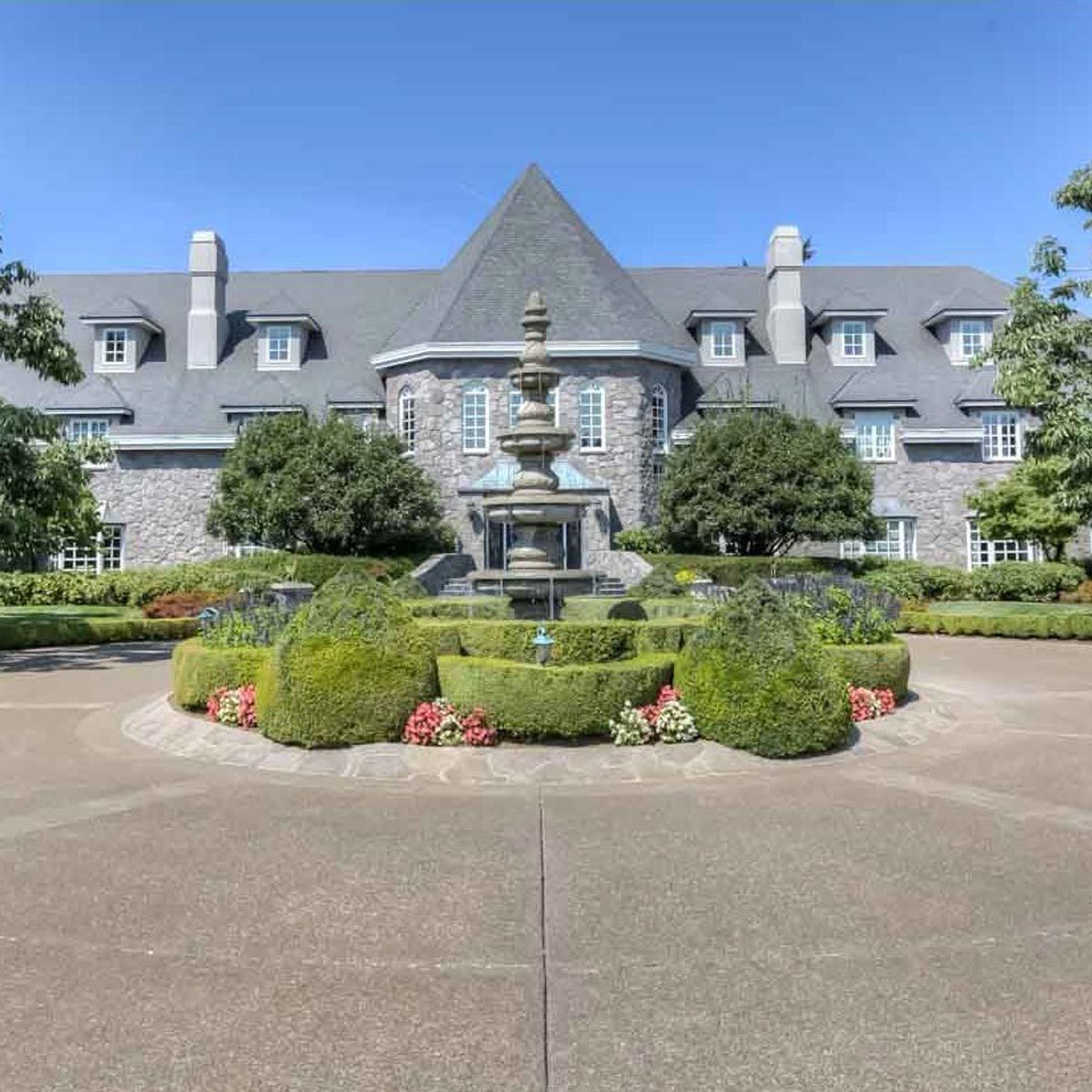 A large stone mansion features a central fountain surrounded by manicured gardens and hedges, set against a clear blue sky.