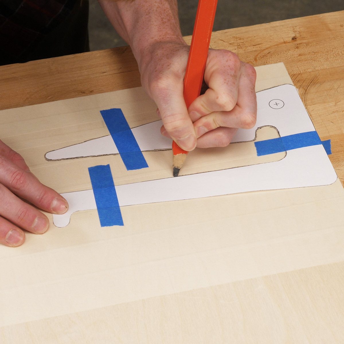 A hand uses a pencil to trace a paper template taped on a wooden surface, preparing for cutting or shaping the material underneath.