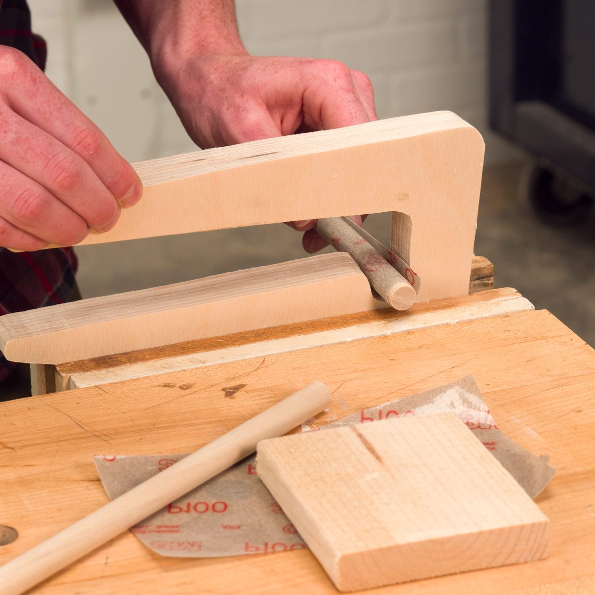 A person uses a tool to shape a wooden dowel, positioned on a workbench with sanding paper and wood pieces nearby, in a workshop.
