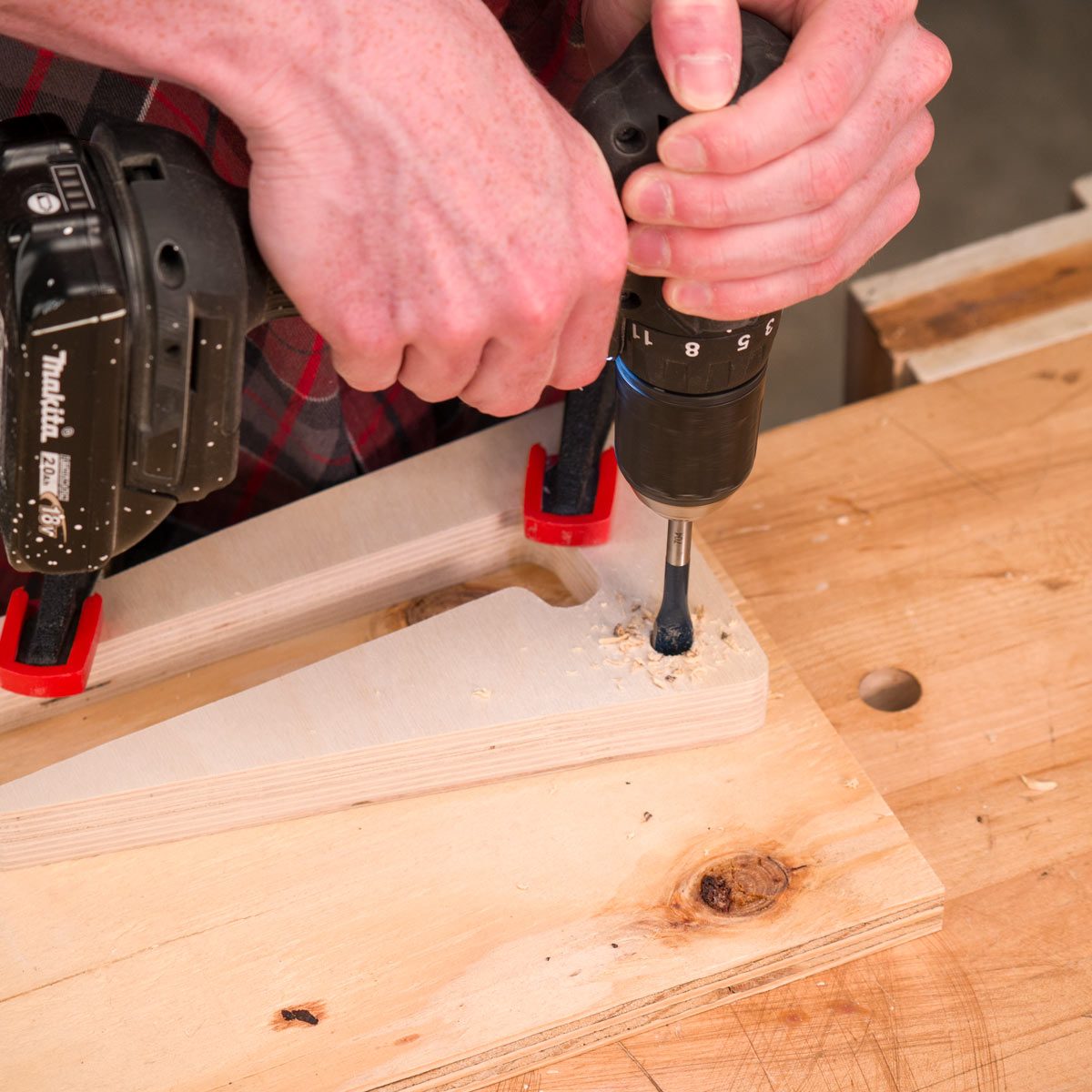 A person drills into a piece of wood, securing the material with clamps. Wood shavings accumulate on the surface, indicating active woodworking.