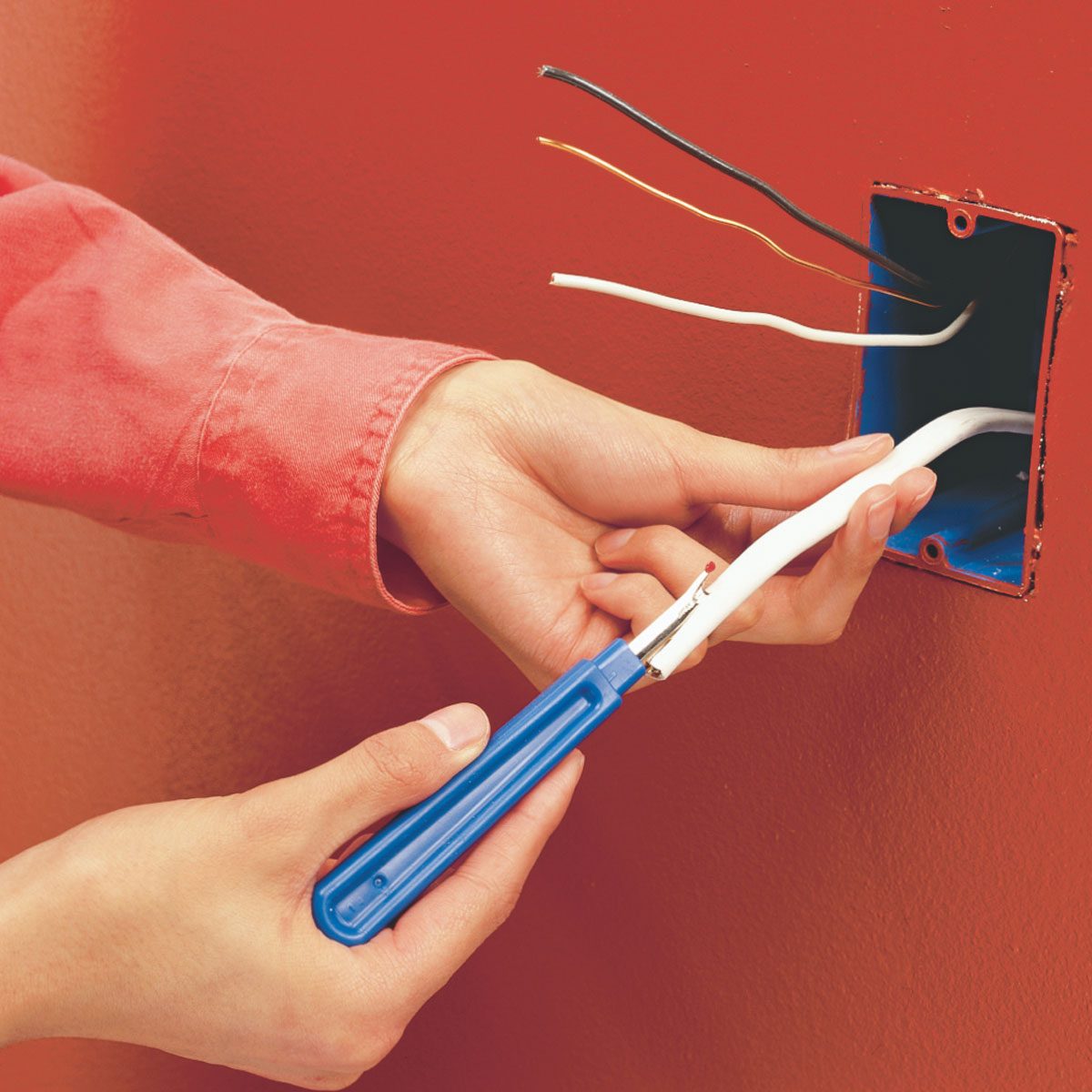 A hand holds a blue wire stripper, stripping insulation from a white wire, while other colored wires emerge from an electrical box on a red wall.