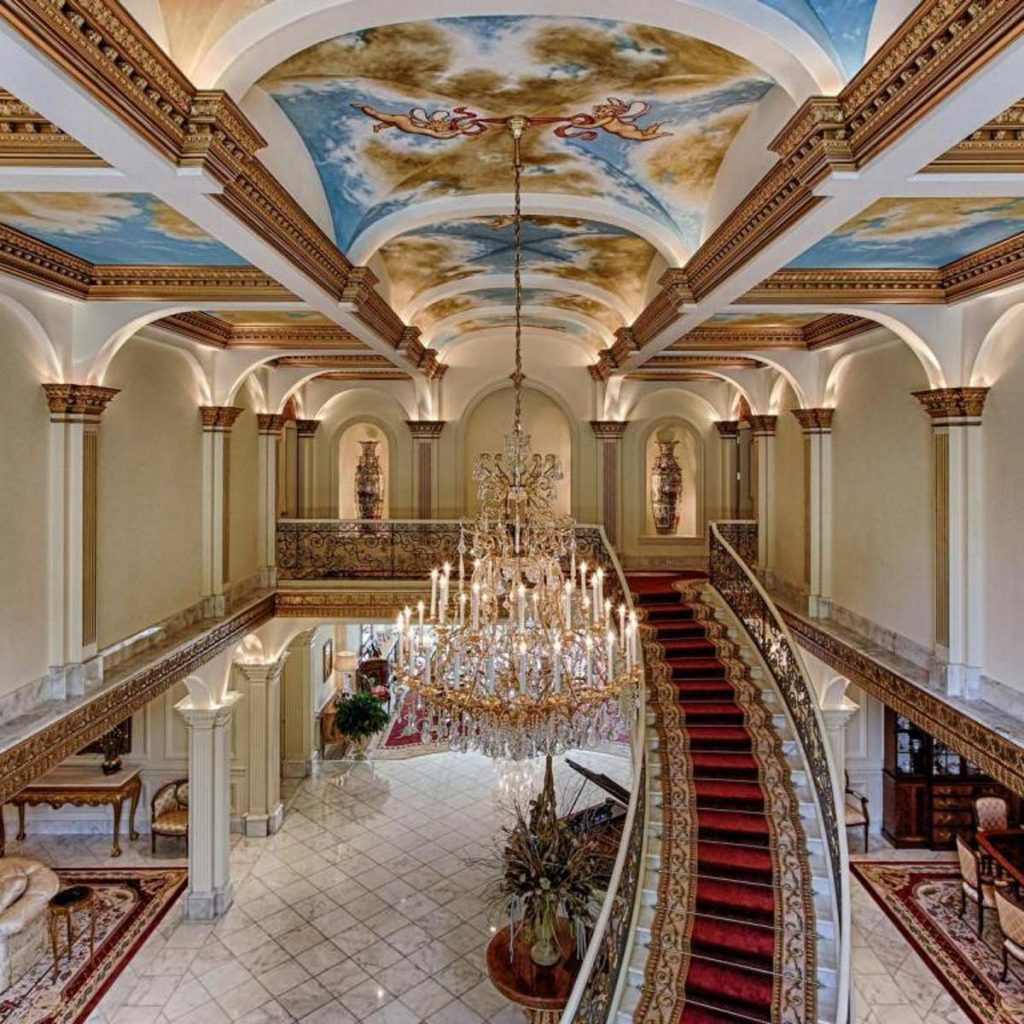 A grand staircase curves upward in an elegant foyer, featuring a shimmering chandelier and ornate decor, illuminated by soft lighting and opulent ceiling artistry.