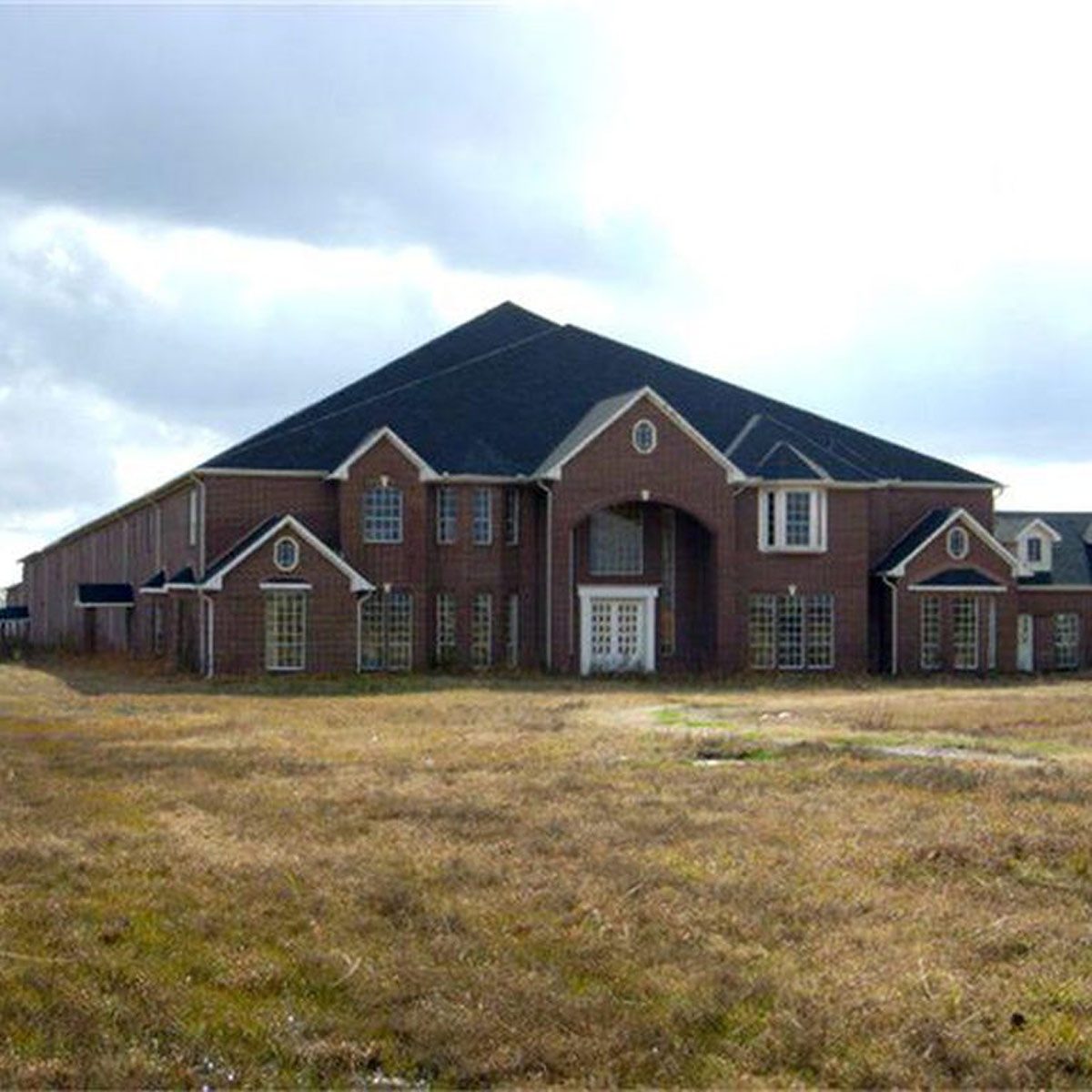 A large, abandoned brick house sits in a grassy field. Its architecture features a peaked roof and multiple windows, under a cloudy sky.
