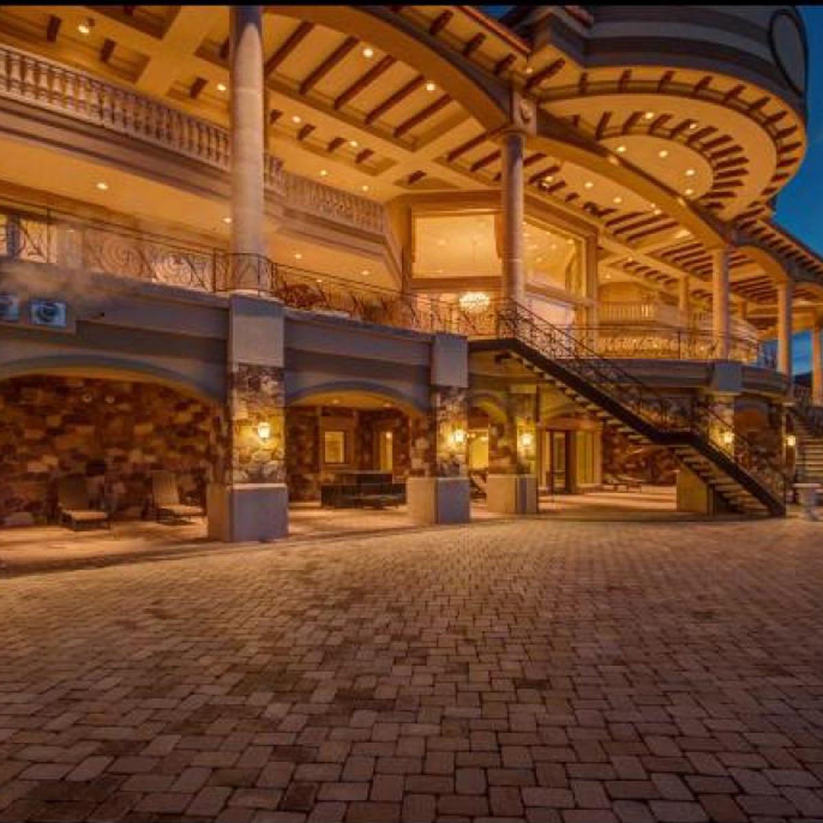A grand multi-level structure with decorative lighting occupies a paved area, featuring stone accents, balconies, and an illuminated entrance, set against a twilight sky.