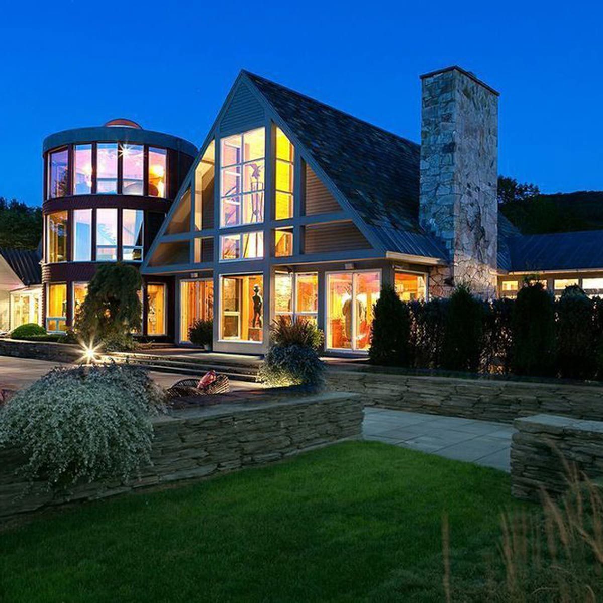 A modern home with large glass windows illuminates the night, showcasing a stone chimney and unique architectural features, surrounded by landscaped greenery.
