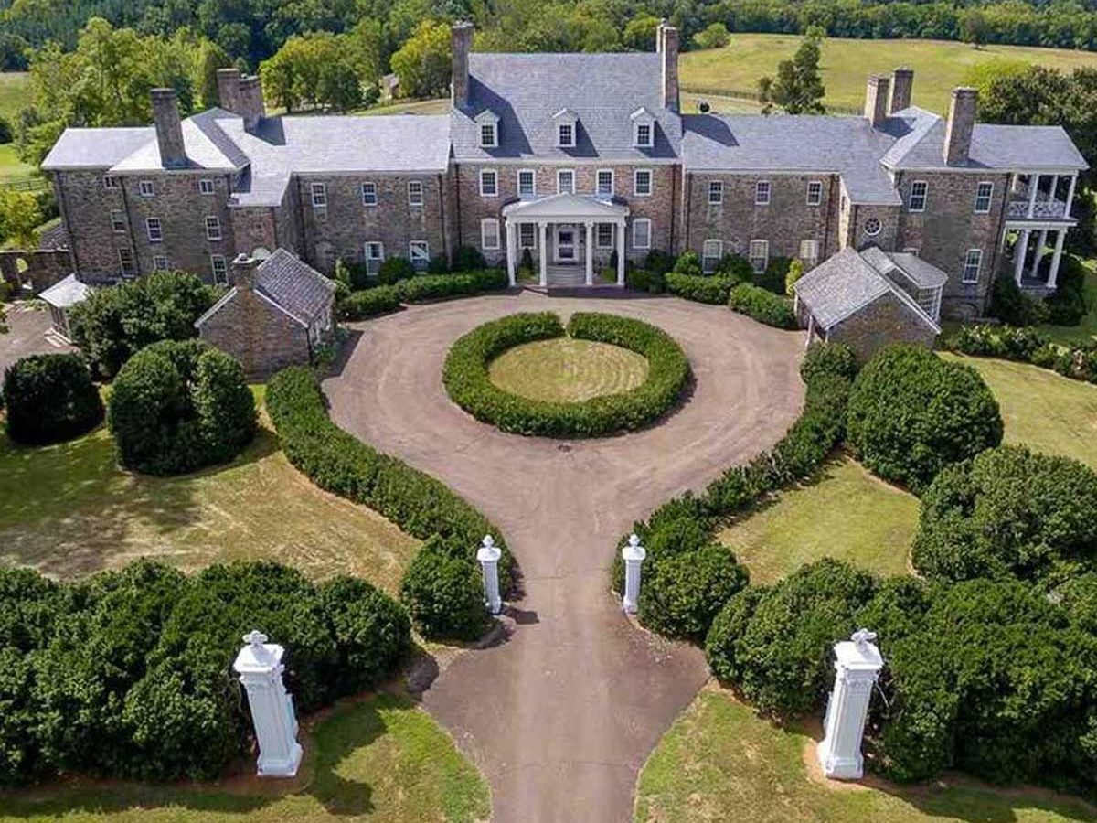 A large stone mansion stands prominently, surrounded by well-manicured lawns and circular hedges, with a spacious circular driveway leading to its entrance.