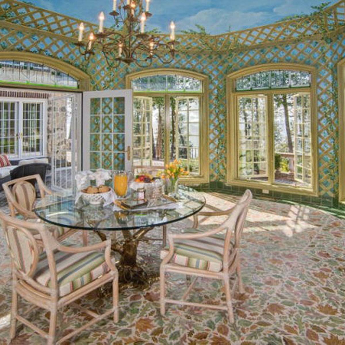 A glass dining table is set with food and drinks, surrounded by plush chairs, in a bright room with large windows and decorative walls.