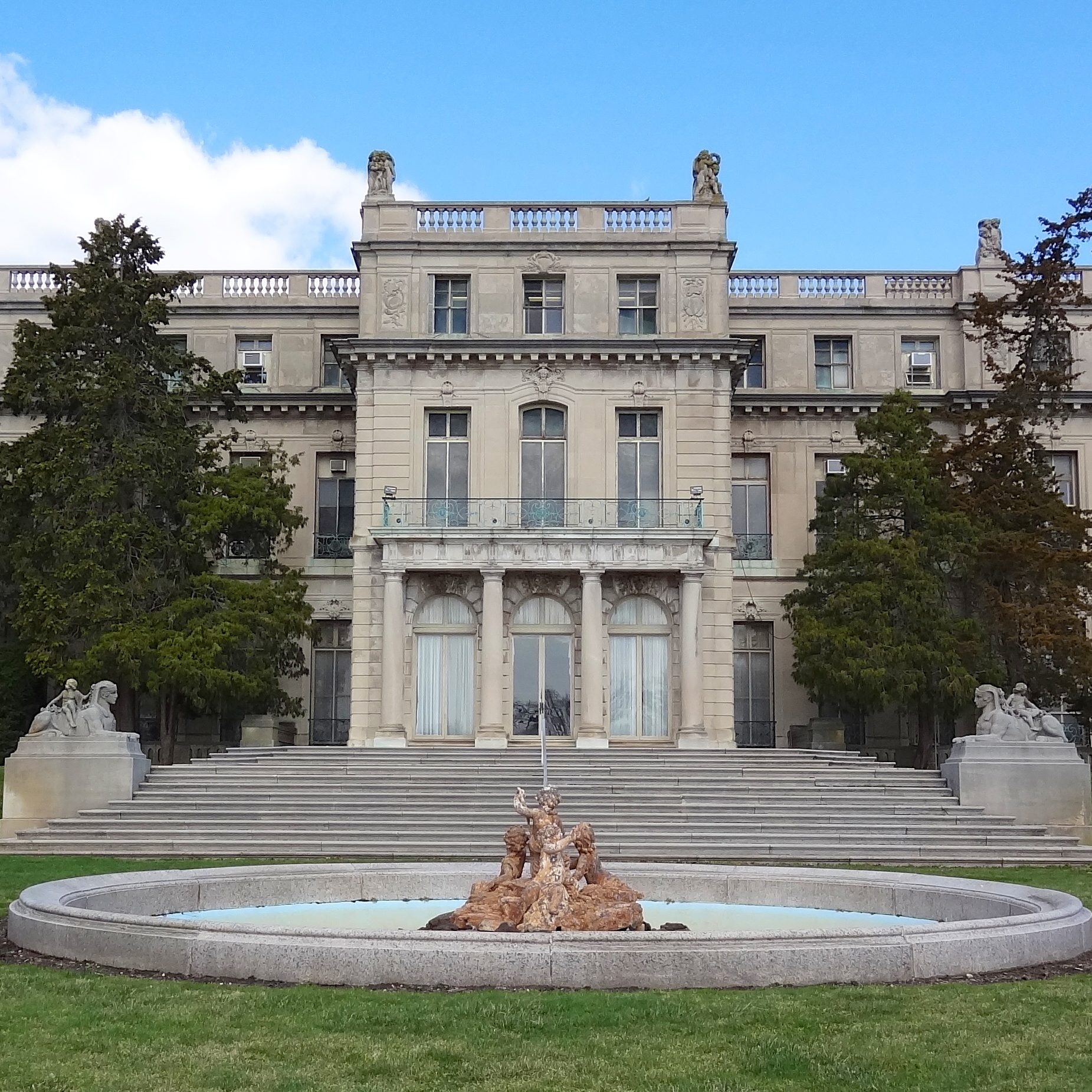 A grand mansion features a central fountain with statues, flanked by trees and steps, set against a blue sky and white clouds.