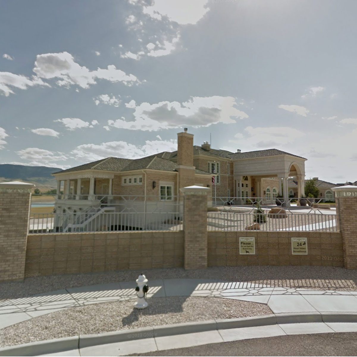 A large, elegant house is situated behind a tall fence, featuring a driveway, decorative landscaping, and a clear blue sky with scattered clouds.