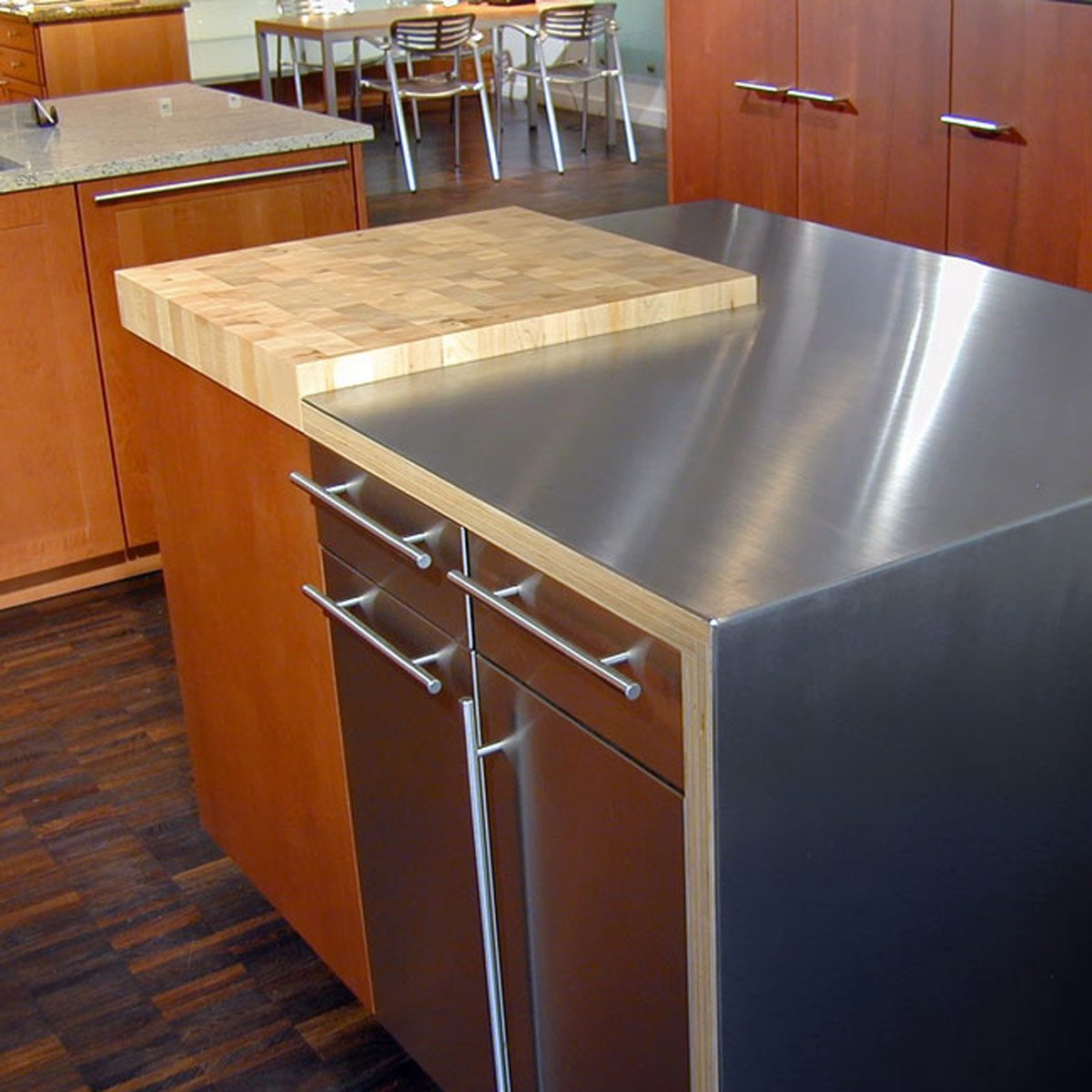 A kitchen island features a stainless steel surface and a wooden chopping block; wooden cabinets and chairs are visible in the background.