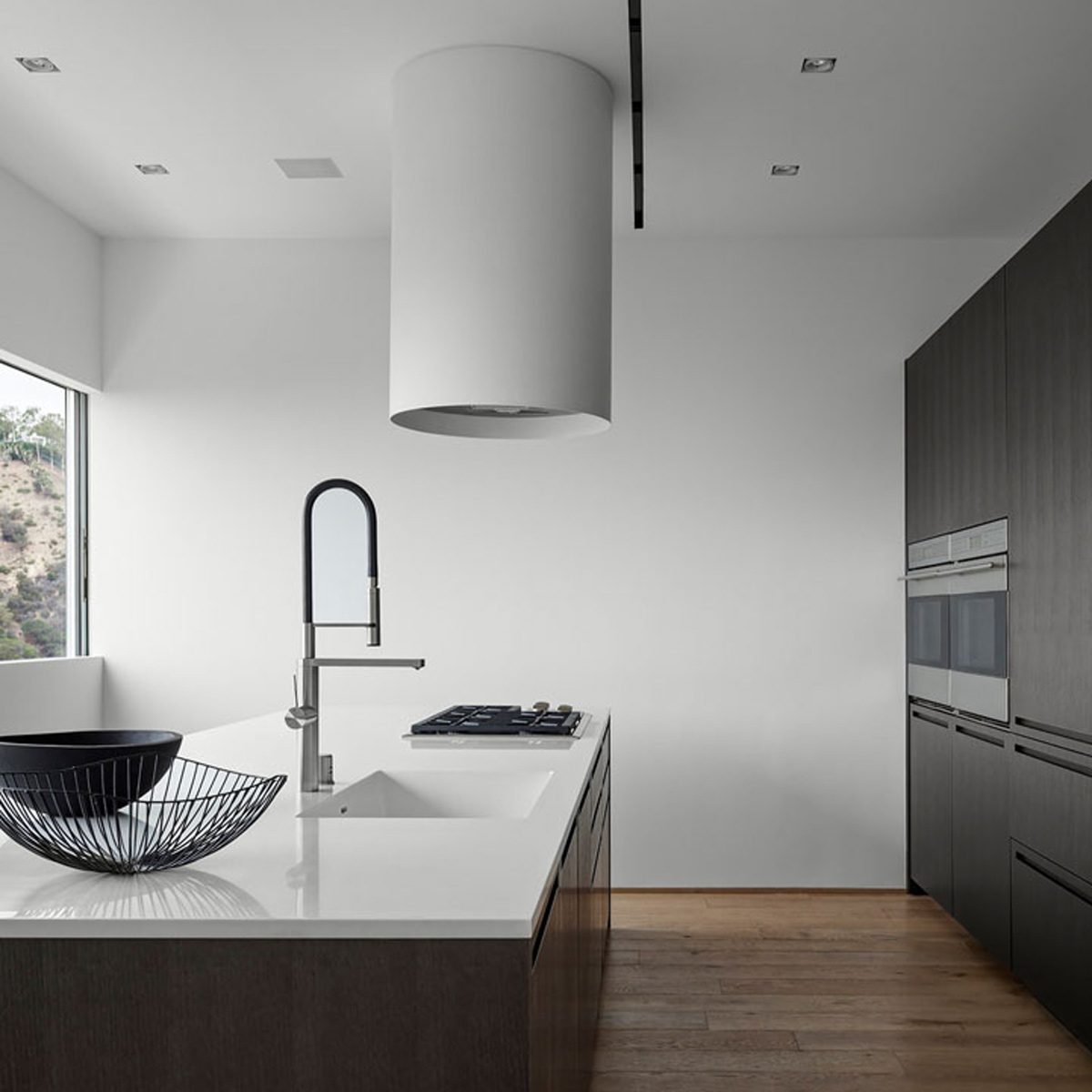 A modern kitchen features a sleek white countertop with a sink, stylish faucet, and a gas stove, surrounded by minimalist cabinetry and a large window.