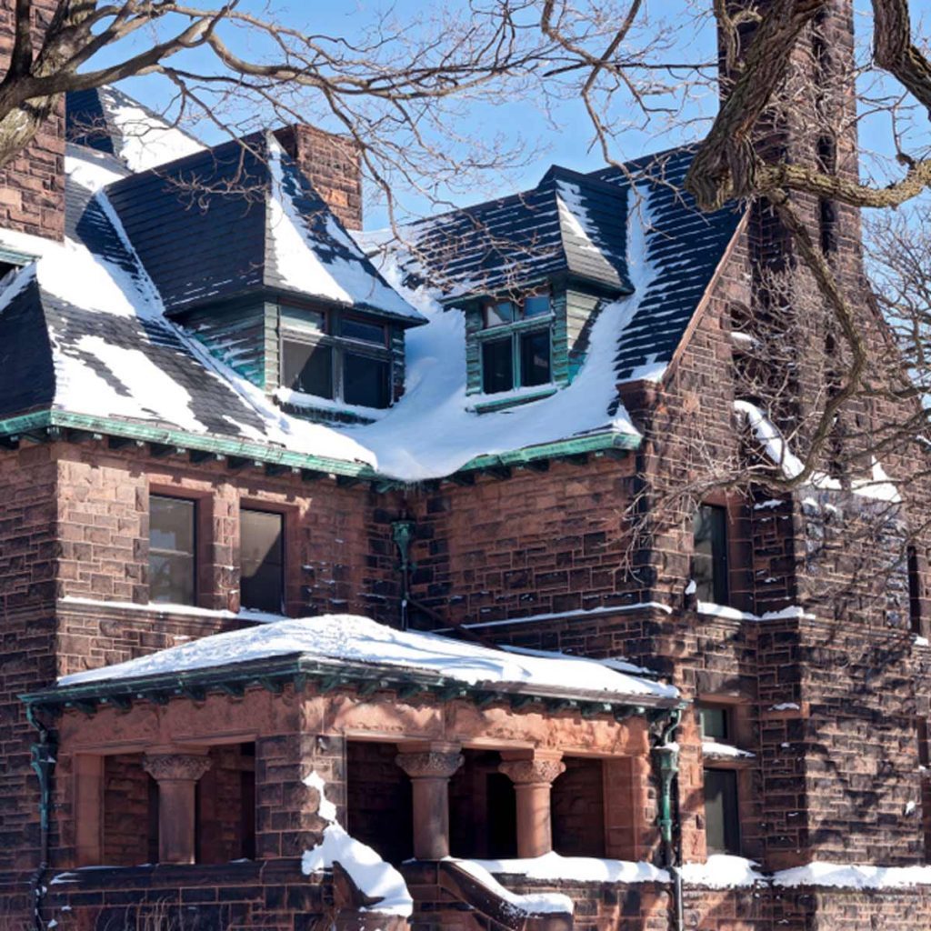 A historic stone house with snow on the roof and a porch supported by columns, surrounded by leafless trees under a clear blue sky.
