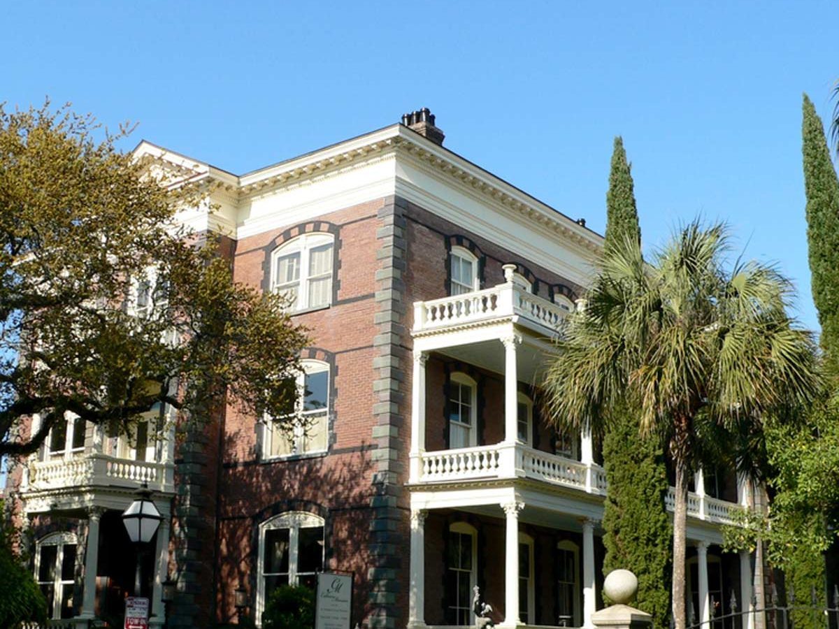 A historic brick building features balconies and decorative railings, surrounded by lush vegetation and palm trees against a clear blue sky.