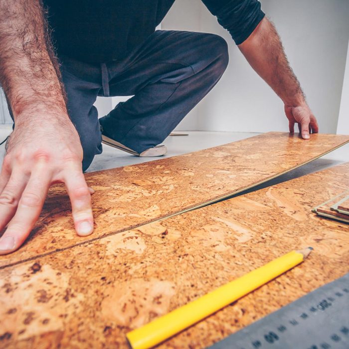 A person kneels, aligning cork flooring planks on a surface, surrounded by tools like a yellow pencil and a ruler in a well-lit room.