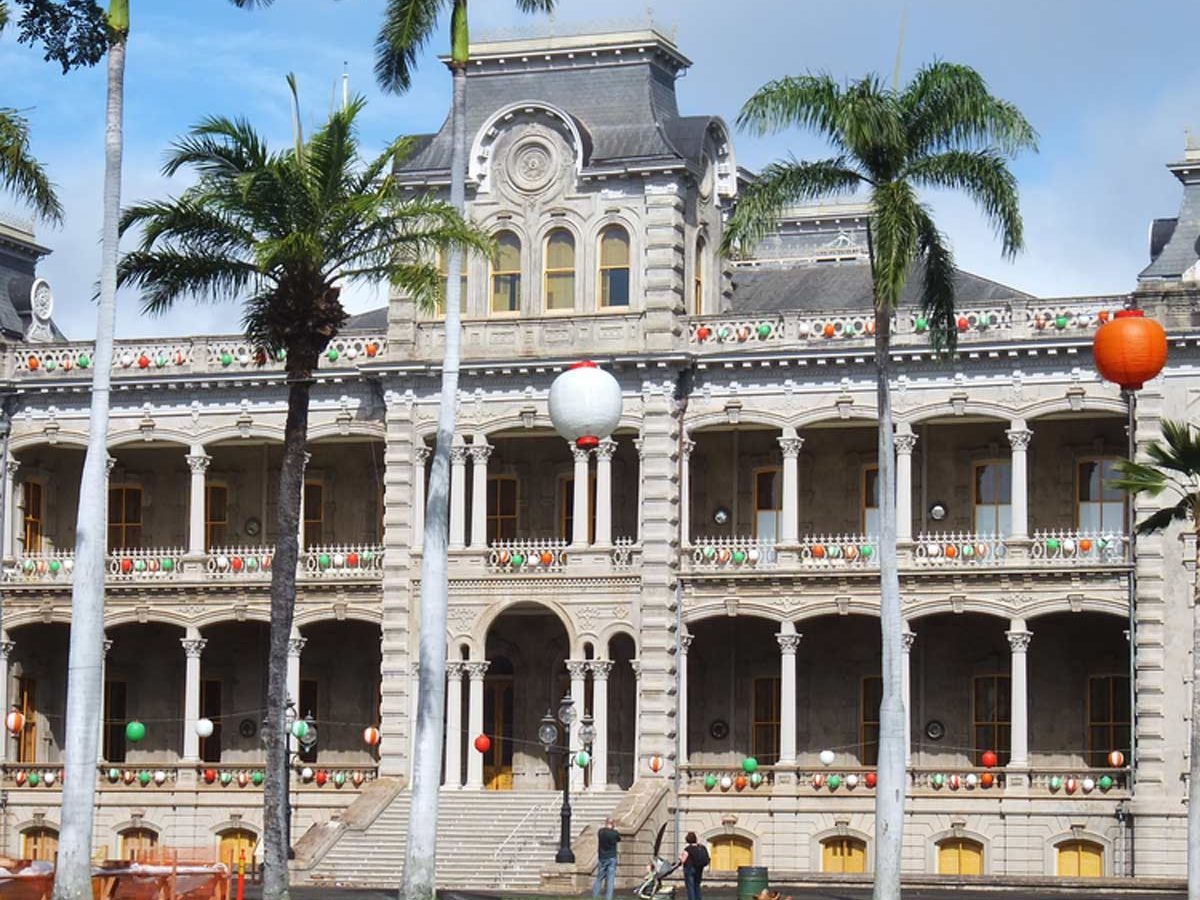 A historic building with large balconies displays decorative lanterns. Two people engage in work on the lawn, surrounded by palm trees and a clear sky.