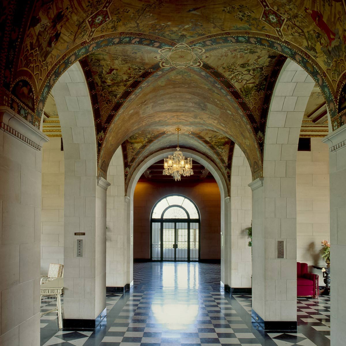 A grand hallway features arched ceilings, ornate decorations, and a chandelier, leading to a large window with doors, enhanced by black and white checkered flooring.