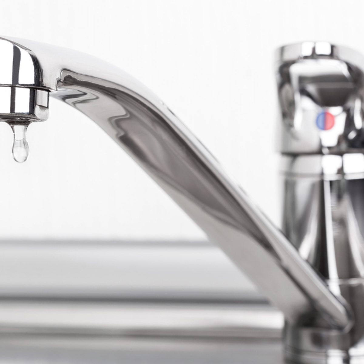 A shiny faucet drips water into a sink, reflecting light. The background is blurred, indicating a simple kitchen or bathroom setting.