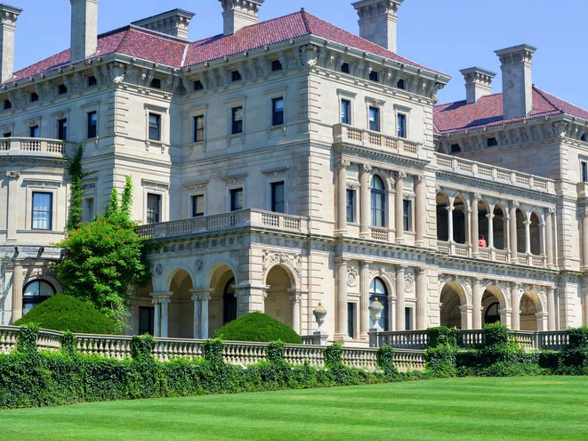 A lavish mansion showcases ornate architecture, with lush green lawns and manicured hedges surrounding it under a clear blue sky.