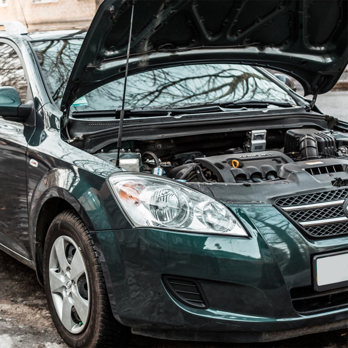 A green car with its hood raised exposes the engine, parked on a street with visible trees and buildings in the background.