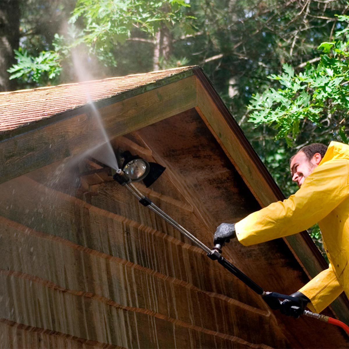 A person in a yellow raincoat uses a pressure washer to clean the roof eaves of a wooden structure surrounded by trees. Water sprays out forcefully.