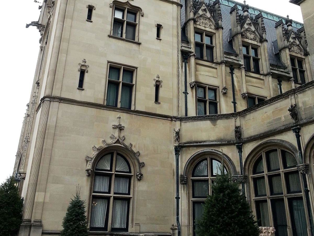 A stone mansion features ornate windows and detailed architecture, with potted evergreen plants at the entrance, set against a cloudy sky.