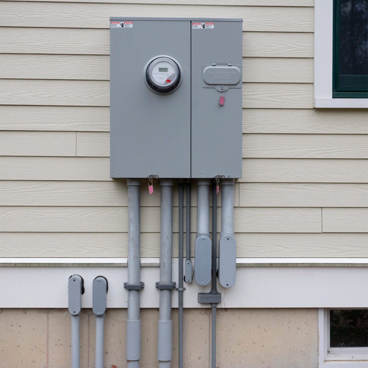 A grey electrical meter and junction box are mounted on a light-colored wall, surrounded by multiple metal conduits leading downward.
