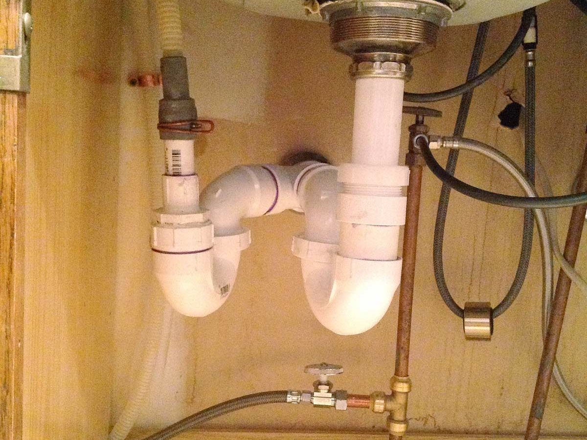 A power panel mounted on a wooden board connects to various metal pipes, situated against a white concrete wall in a basement environment.