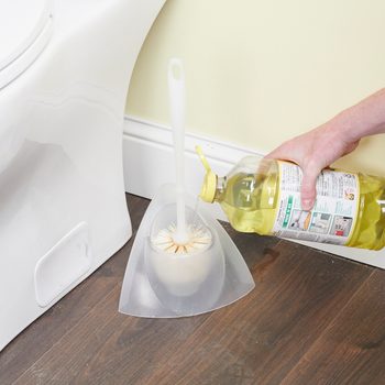 A hand pours cleaning liquid onto a toilet brush resting in a holder beside a toilet, set against a light-colored wall and wooden floor.