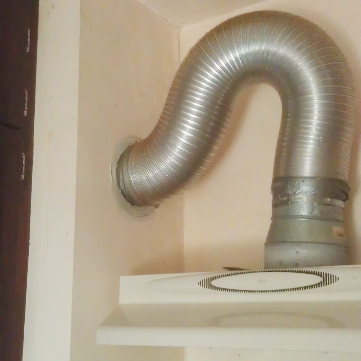 A silver flexible duct curves from a wall vent, connecting to a metallic fitting. The surrounding area features a cream-colored wall and a white shelf beneath.
